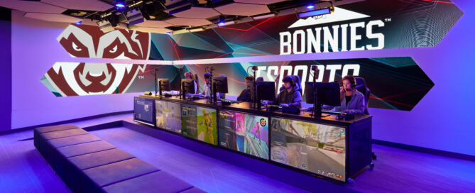 Completed competitive esports arena at St. Bonaventure University featuring a dedicated team competition stage, professional PC gaming stations, integrated broadcast displays, acoustic ceiling panels, and branded graphic walls with dynamic lighting.