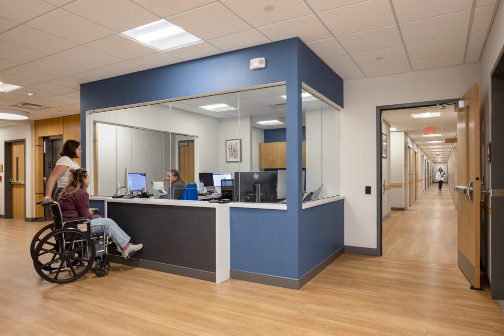 A person in a wheelchair checks in at a reception desk at the GLOW YMCA, and a doctor approaches down the hall.