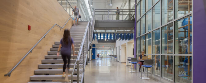A spacious hallway inside the GLOW YMCA; people sit at tables, walk across an elevated walkway, or climb the stairs to the second floor.
