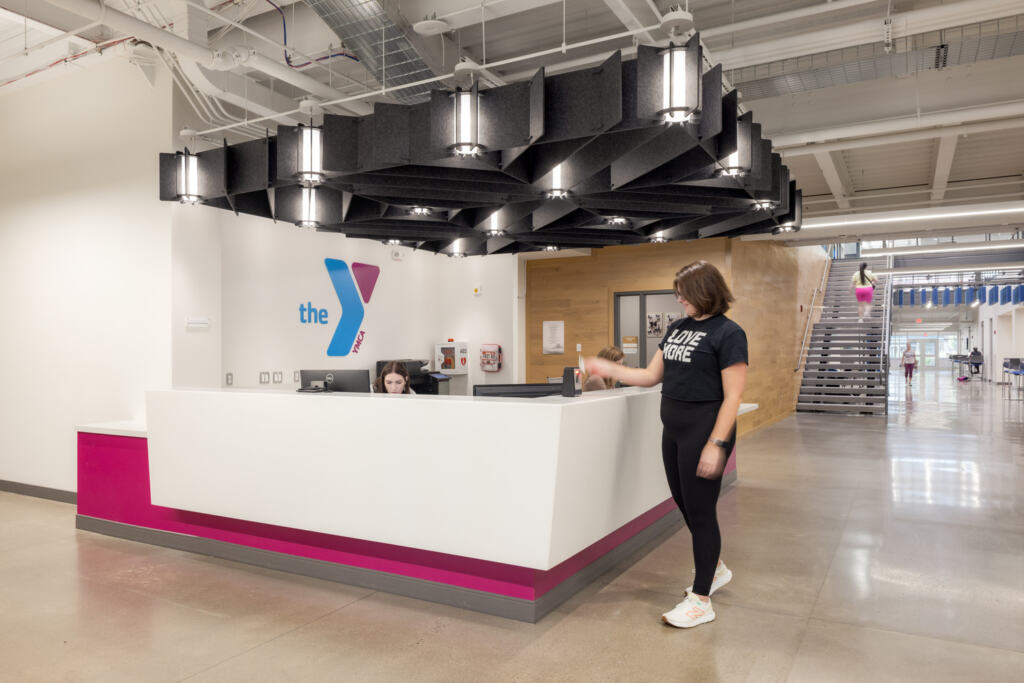 A woman signs in at the GLOW YMCA Healthy Living Campus's front desk with a "Y" logo decal on the wall.
