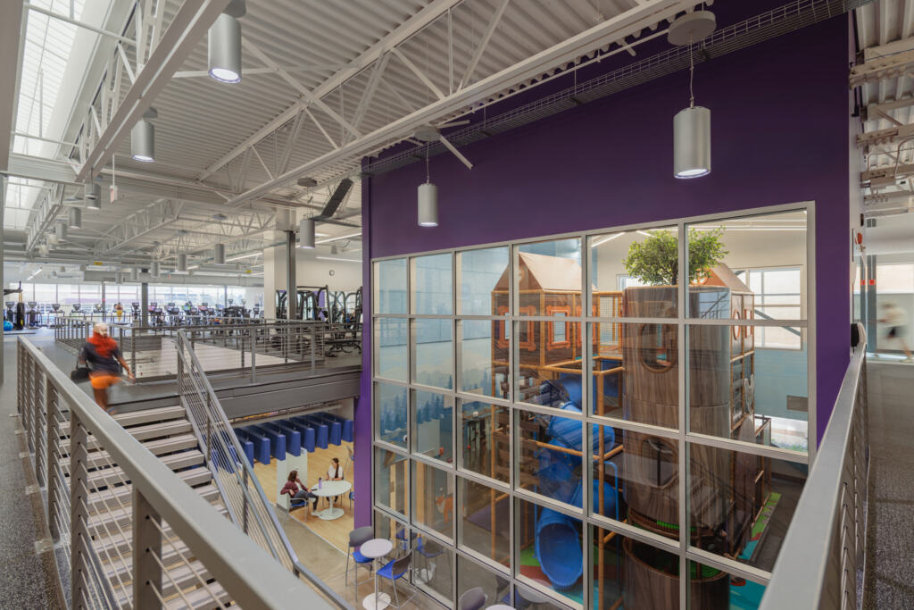 The Kid's Adventure Space, a colorful forest-themed playground, through tall windows and a purple wall inside the GLOW YMCA.