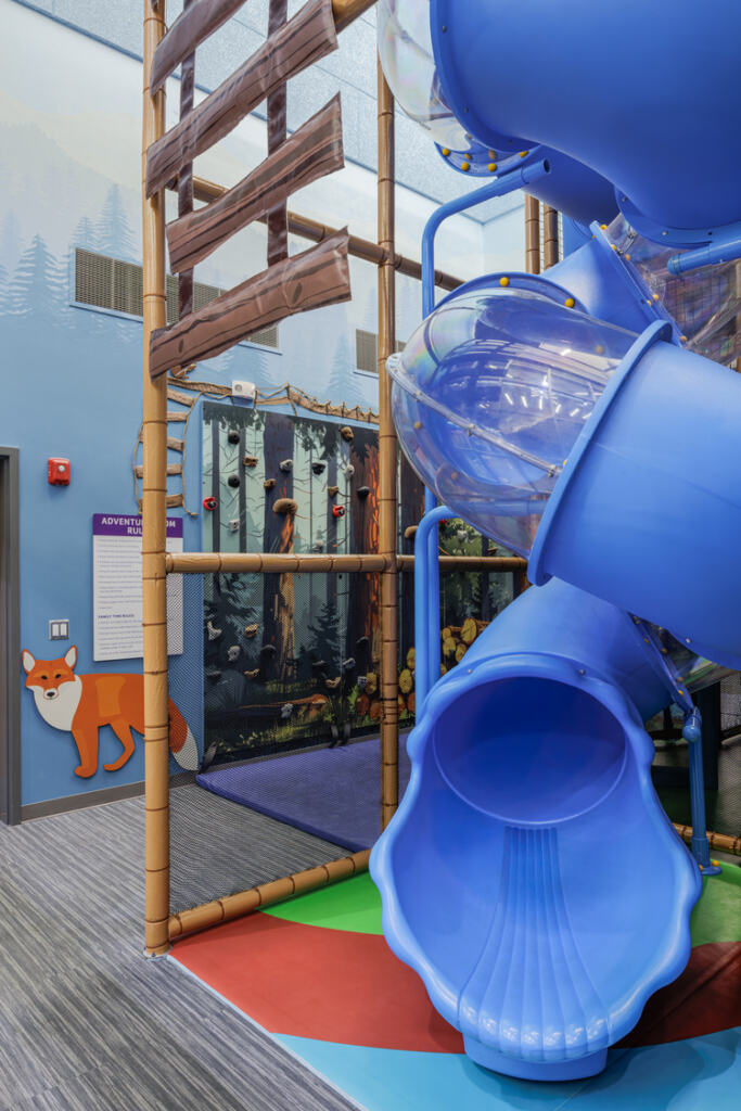 A swirly blue slide and a forest-themed climbing wall inside the colorful GLOW YMCA Kid's Adventure Space.