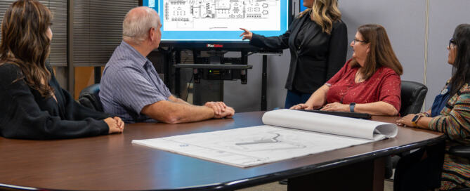 A woman is pointing to a school floor plan while standing in front of people seated at a table as part of CPL's on-call K-12 services.