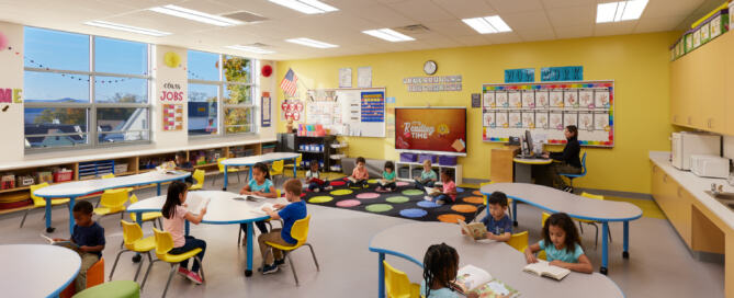 An Ossining UFSD Park Elementary School classroom has vibrant yellow walls, colorful decorations, and a dozen young children independently reading at tables and on the rug.