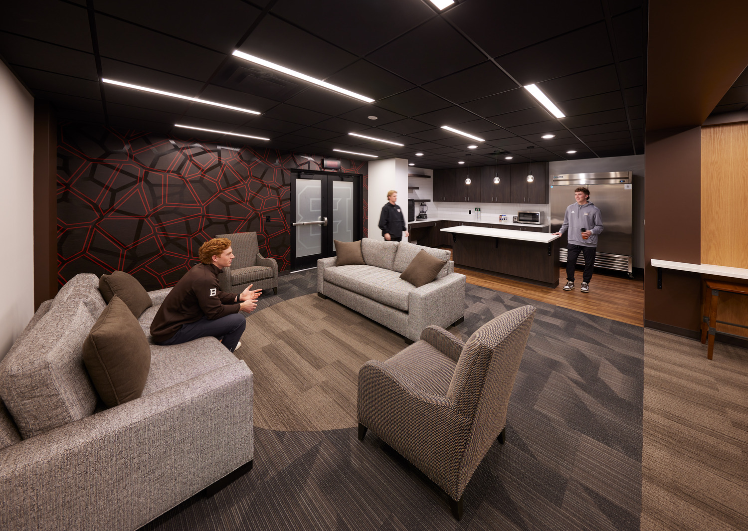 A common area in St. Bonaventure's new locker room with grey couches, a kitchenette, and a patterned accent wall.