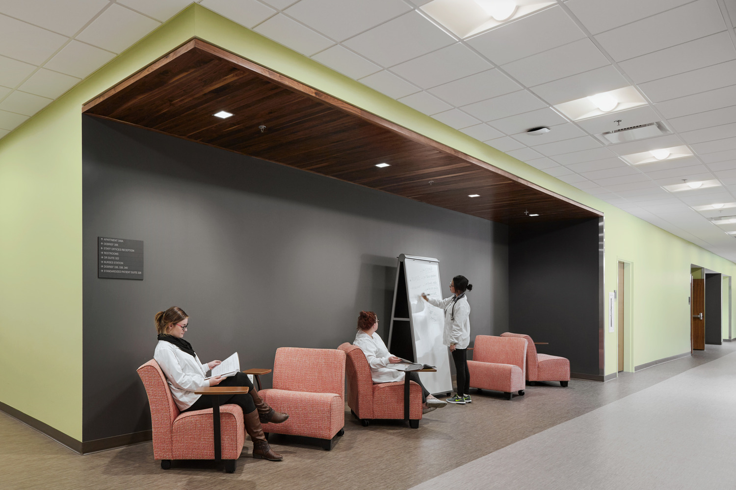 Modern hallway with three people in lab coats, two seated on salmon colored chairs reading, one standing and writing on a whiteboard at Union Square Campus. Sleek, minimalist design.