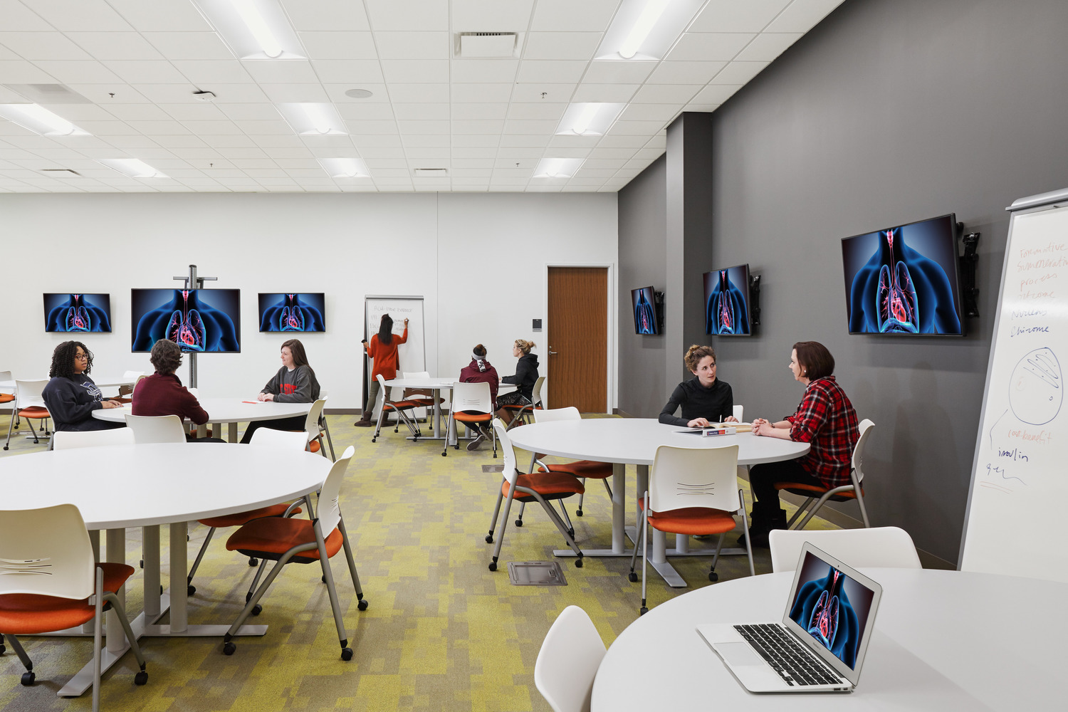 A modern classroom with round tables, occupied by groups of students engaged in discussions. Screens display anatomical images, and one person writes on a whiteboard at Union Square Campus.
