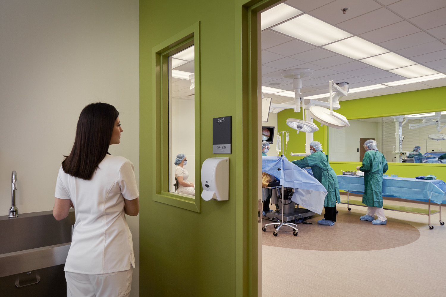 A woman in white scrubs stands outside a brightly lit operating room, observing through a window. Inside, surgeons in green scrubs perform a procedure at Union Square Campus.