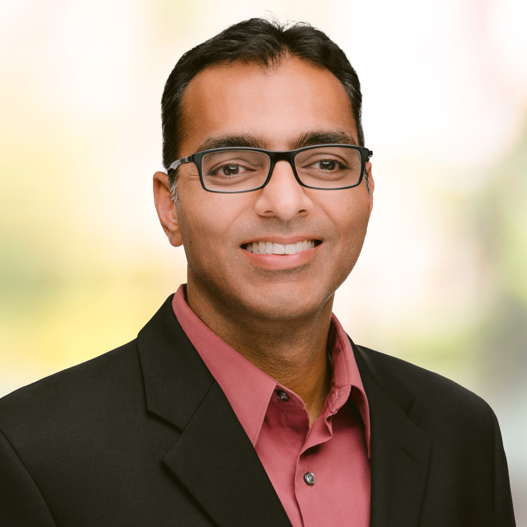 Smiling man with black hair and glasses in salmon colored shirt and black suit jacket