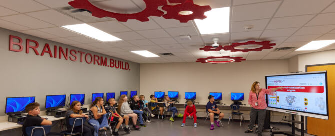 A modernized K-12 facility classroom at the Bolivar / Richburg Central School District with children seated in a semicircle facing a teacher, who is presenting on a screen. Red gear decorations hang from the ceiling, and "BRAINSTORM. BUILD." is written on the wall. The room conveys a creative, educational atmosphere.