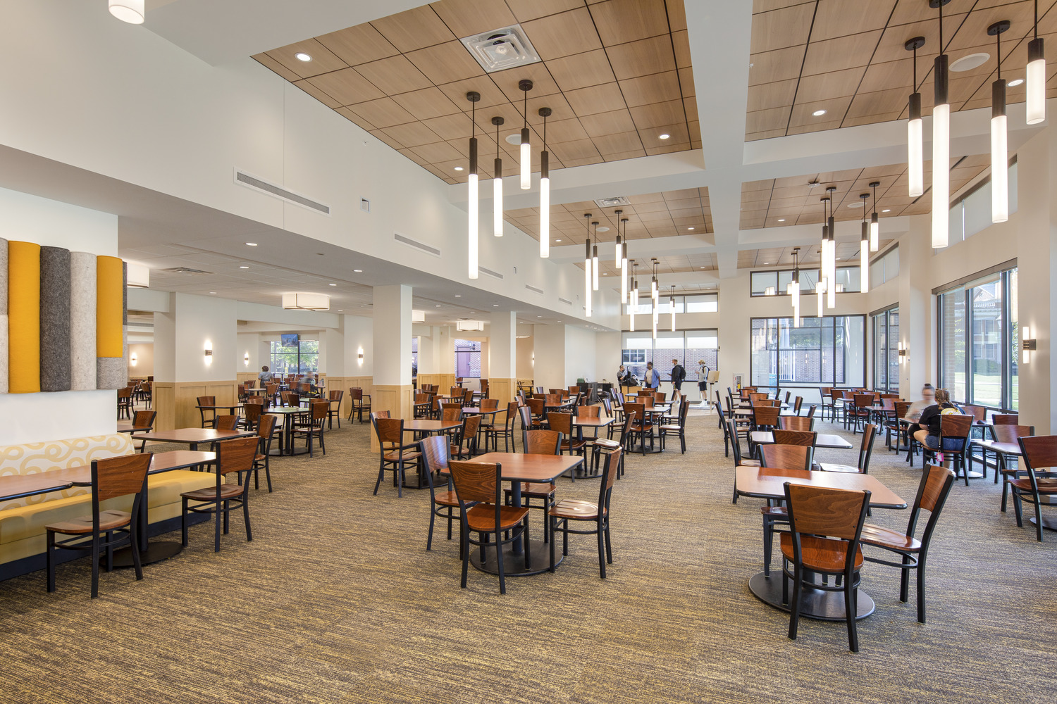 Image shows a modern dining hall with hanging lighting and many tables surrounded by chairs. Students are sitting at the tables together and eating food. This is the Randolph-Macon College Estes Dining Hall.