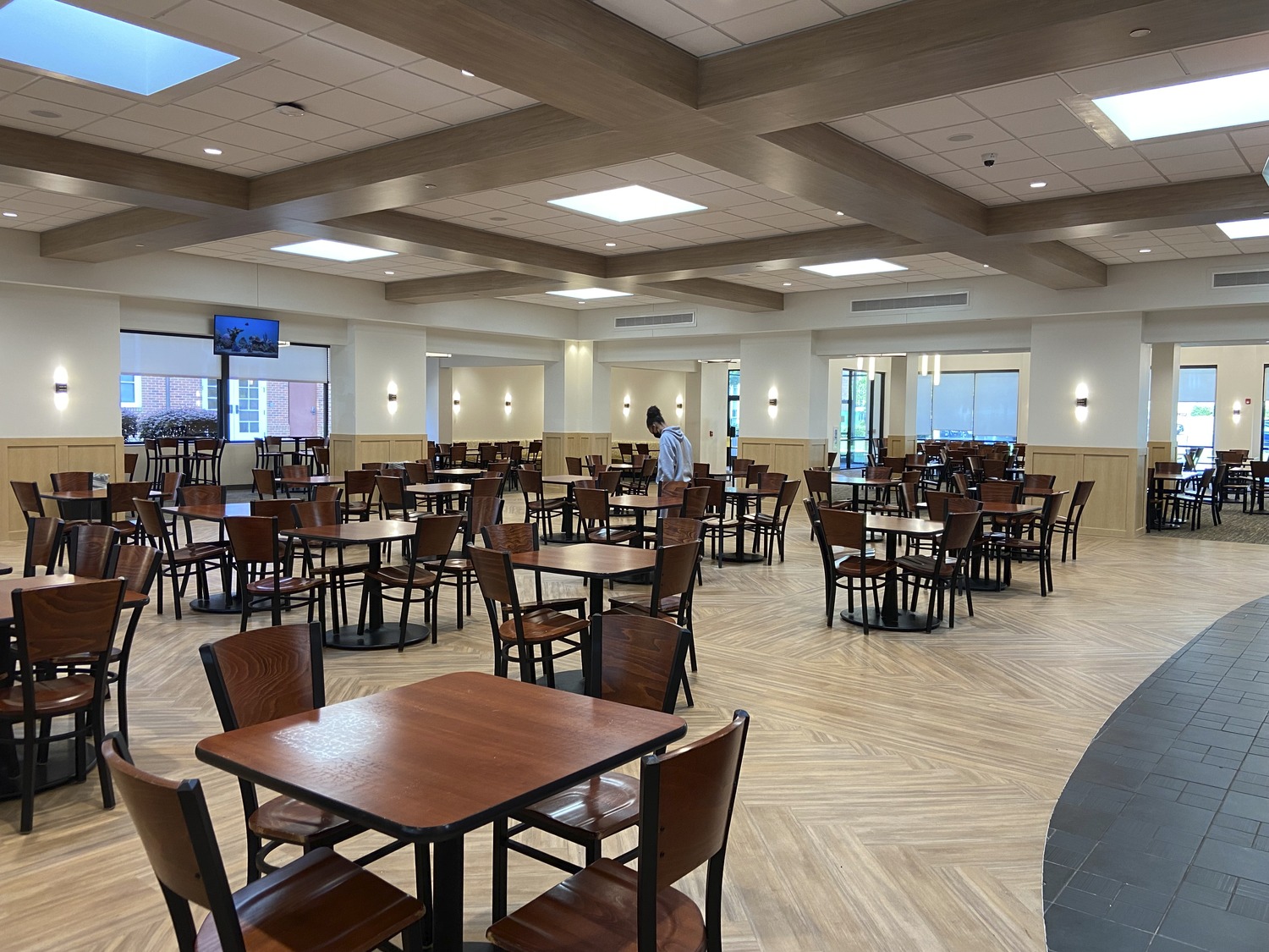 Image shows a modern dining hall with hanging lighting and many tables surrounded by chairs. Students is standing at a table. This is the Randolph-Macon College Estes Dining Hall.