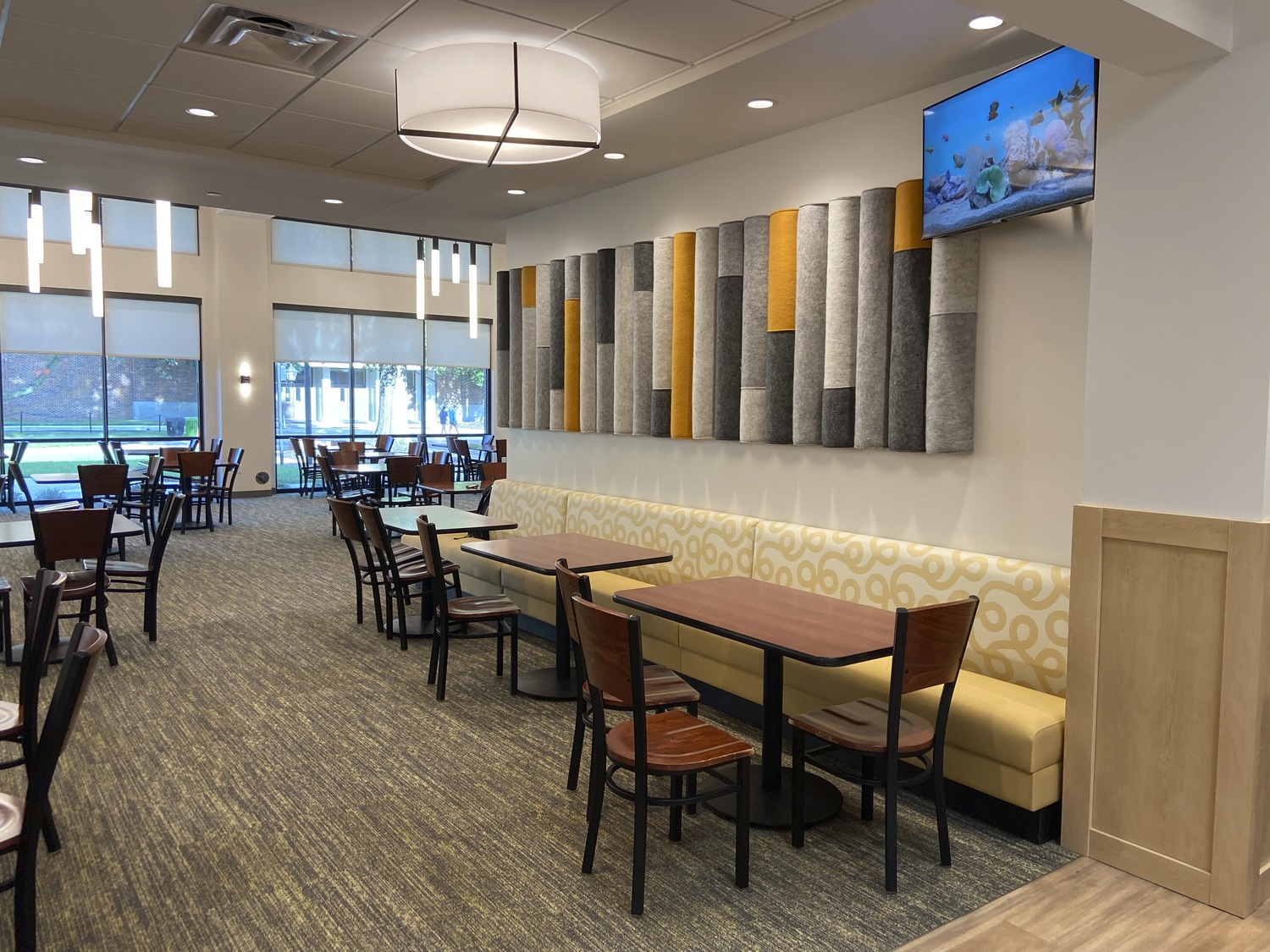 Image shows a modern dining hall with hanging lighting and many tables surrounded by chairs. This is the Randolph-Macon College Estes Dining Hall.