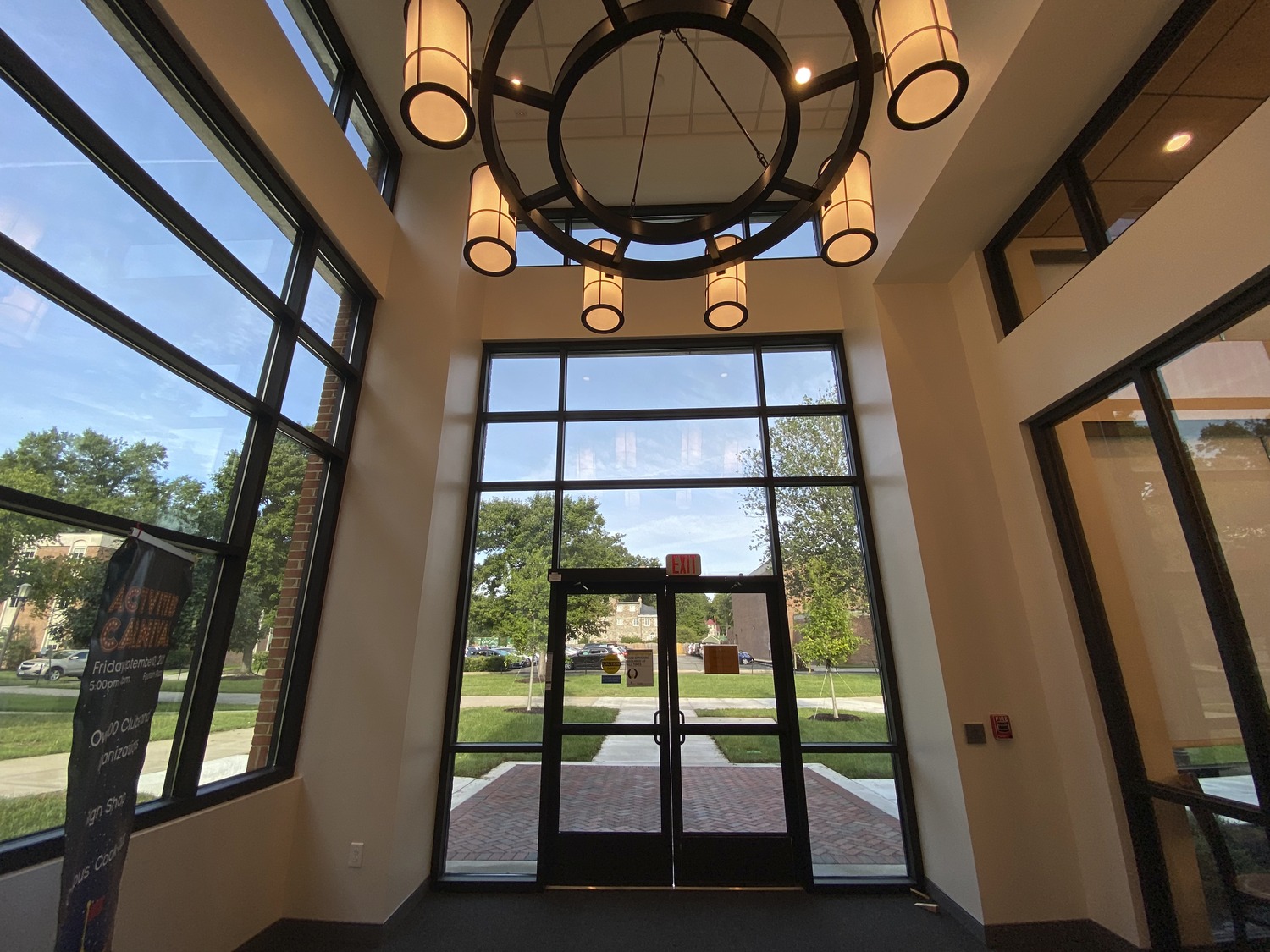 Spacious modern lobby with tall glass windows, circular chandelier overhead. Outside view shows green trees and brick pathway. Bright and inviting atmosphere at Randolph-Macon College Estes Dining Hall.
