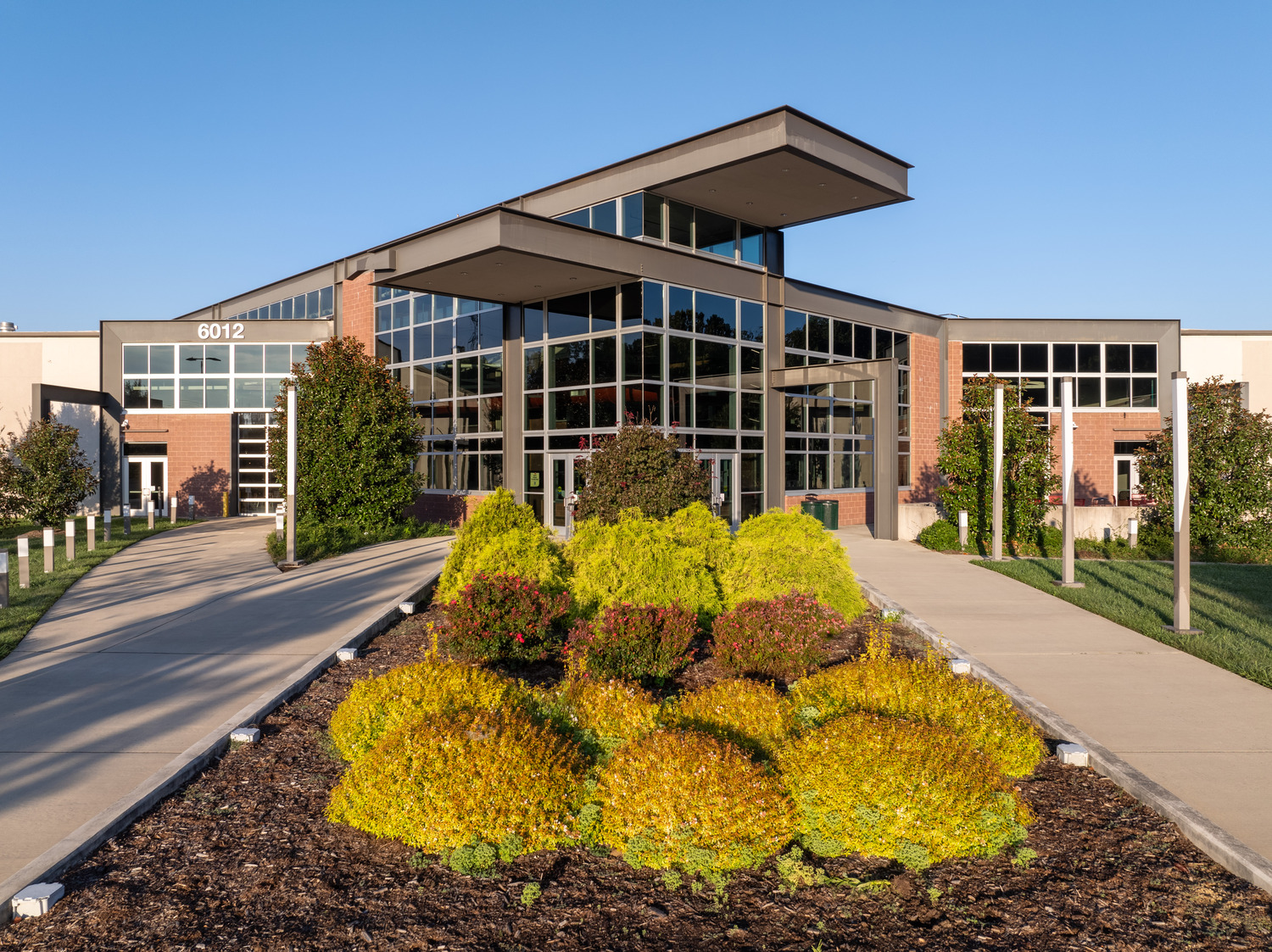 R17_50048_00_GTCC_medium Modern building with large glass windows and a red brick facade. A landscaped garden with vibrant green shrubs lines the entrance pathway under clear blue skies at GTCC.