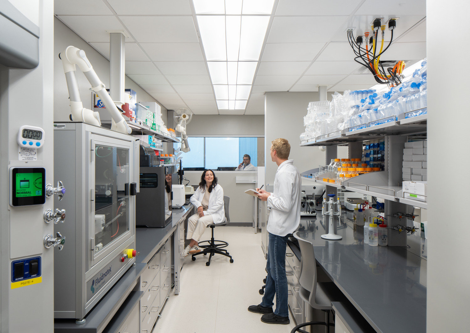 R24_16126_00_USF_ISA_Lab_N2_medium A modern lab with bright lighting, featuring a man and woman in lab coats. They are engaged in conversation, surrounded by lab equipment and shelves of bottles at USF - ISA Biomedical Labs.