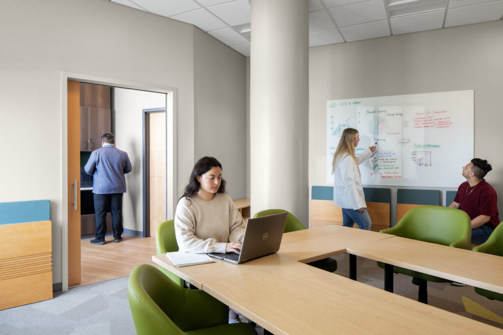 R24_16126_00_USF_ISA_Lab_N4_medium A modern space at USF - ISA Biomedical Labs, with four people: one working on a laptop, another writing on a whiteboard, and a person watching. A fourth is in a kitchenette.