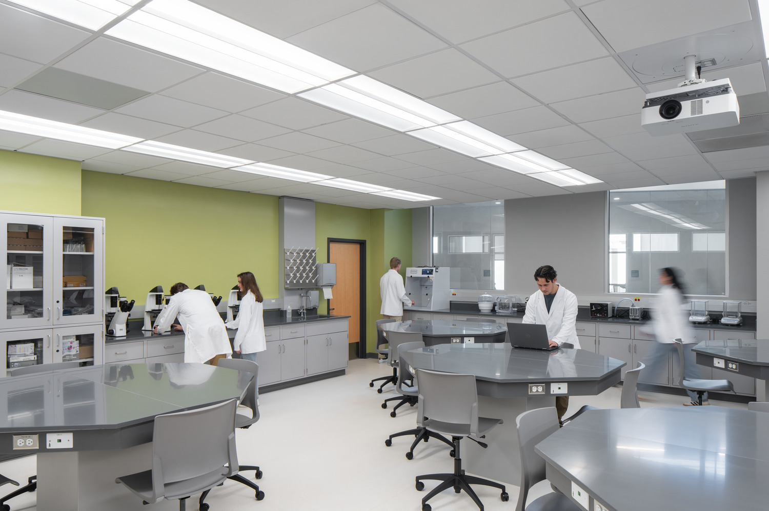 R24_16126_00_USF_ISA_Lab_N5_medium A modern laboratory at USF - ISA Biomedical Labs, with four people in white lab coats conducting experiments and using laptops. The room is bright with gray workstations and green walls.