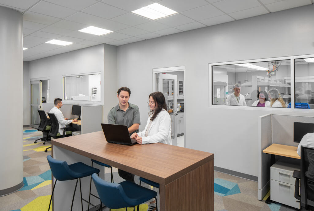 R24_16126_00_USF_ISA_Lab_N7_medium Modern office with a collaborative space where a man and woman work on a laptop at a high table. Others work at desks and behind glass windows at USF - ISA Biomedical Labs.