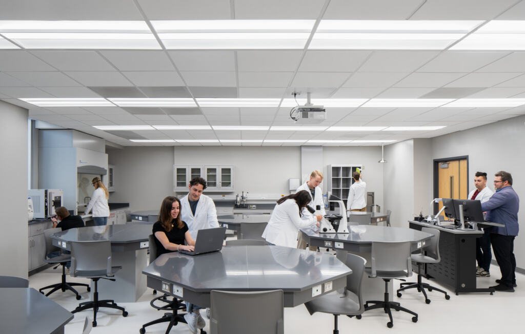 A modern space at USF - ISA Biomedical Labs, with several students in lab coats working around large gray tables. Some use computers, while others engage in discussion, conveying focus and collaboration.