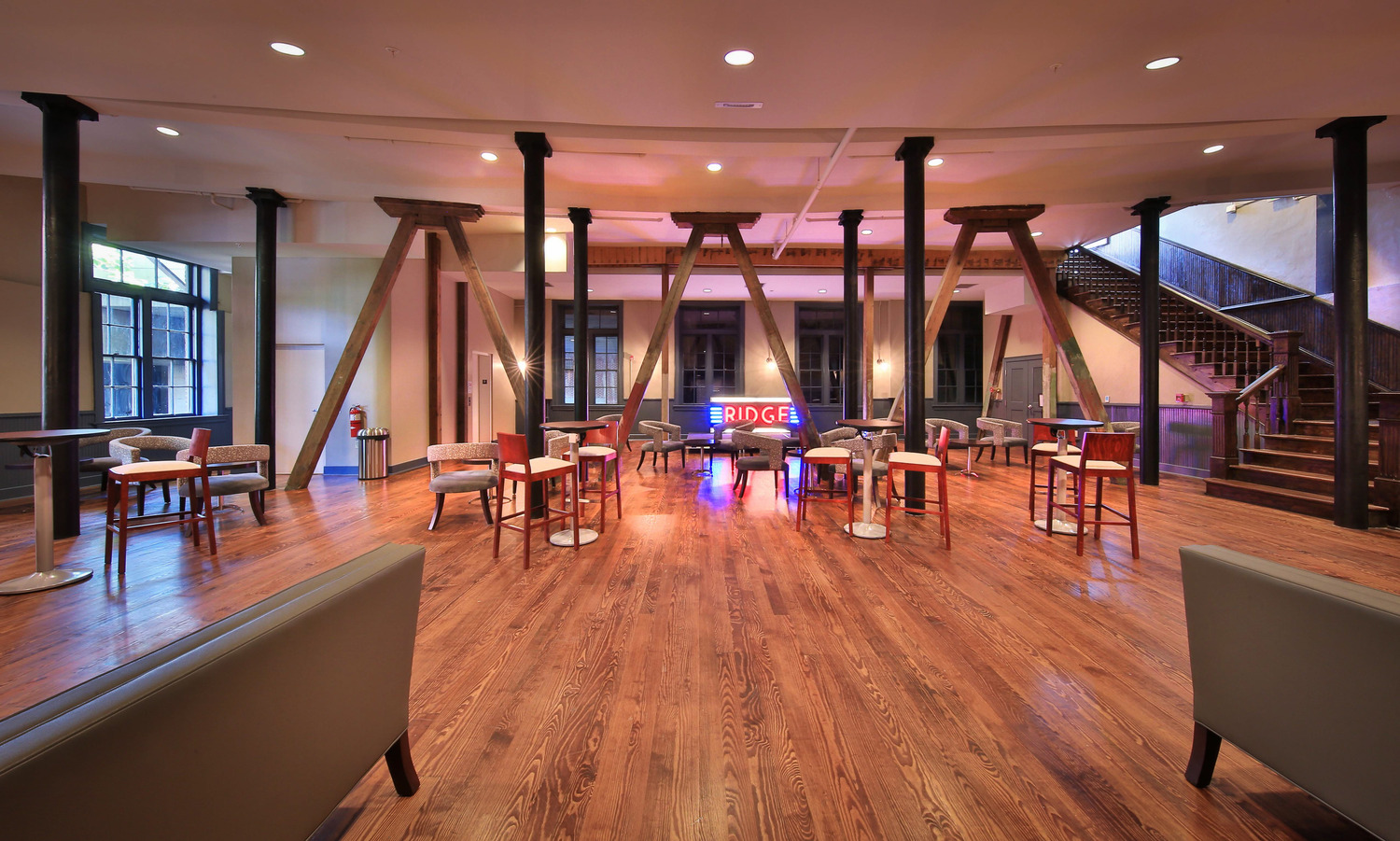 Spacious room with wooden floors, modern furniture, and a neon sign reading "BRIDGE." High ceilings and large windows create an airy, welcoming atmosphere.