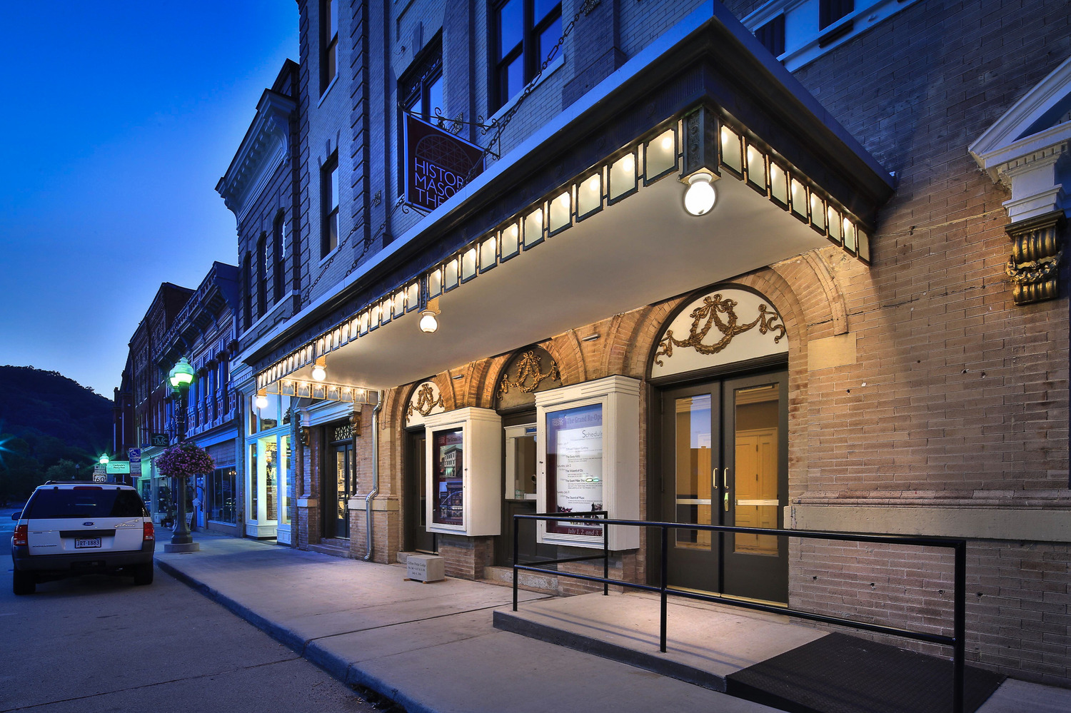 Historic Masonic Theatre with illuminated marquee at dusk, displaying vintage charm. Nearby street features old-fashioned buildings and a parked car.