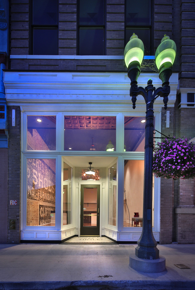 Street view of a vintage storefront with illuminated windows and a hanging light. A lamppost with glowing green lights and pink flowers adds charm.