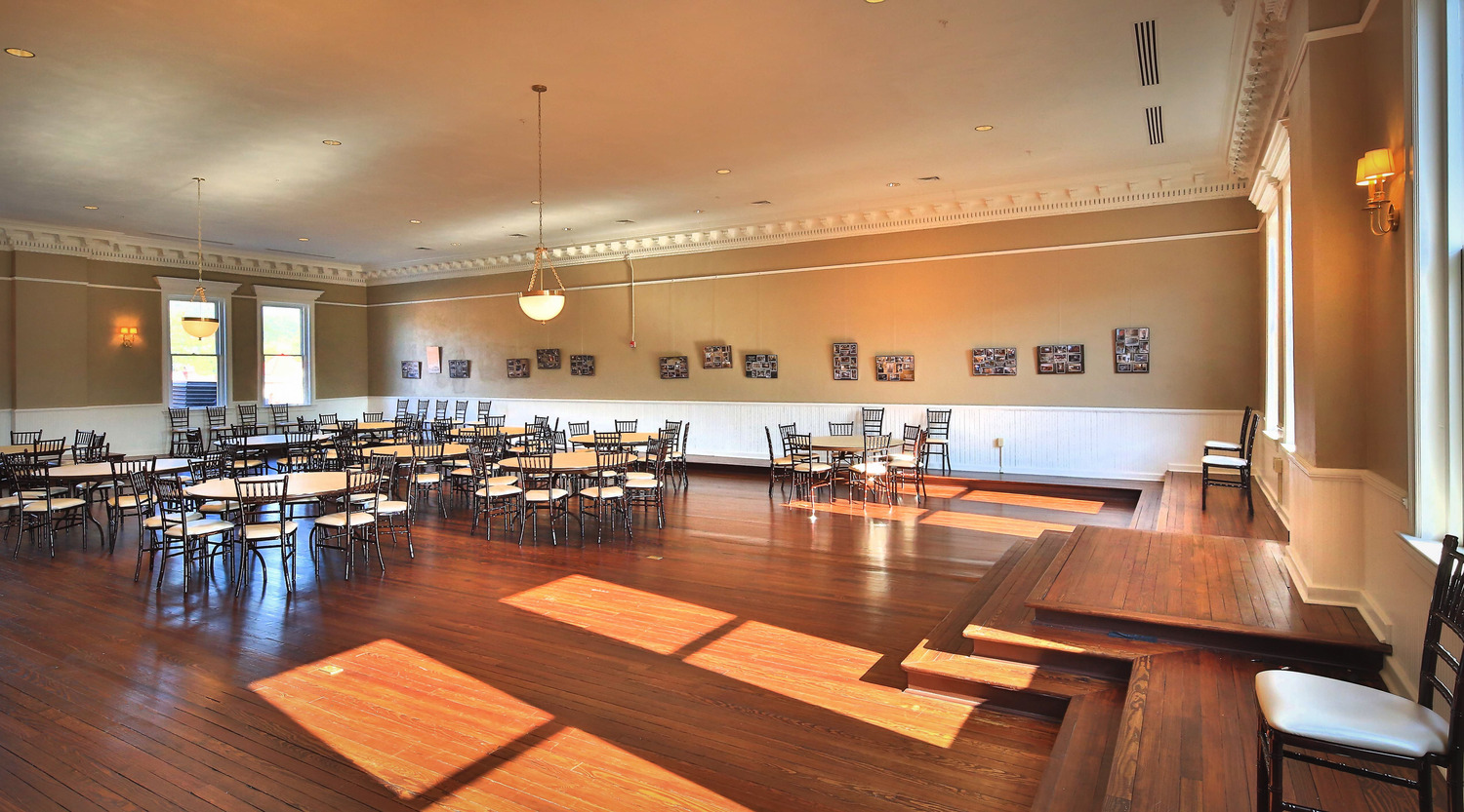 Spacious, sunlit banquet hall with wooden floors, round tables, and chairs. Soft lighting, large windows, and framed artwork create an elegant ambiance.