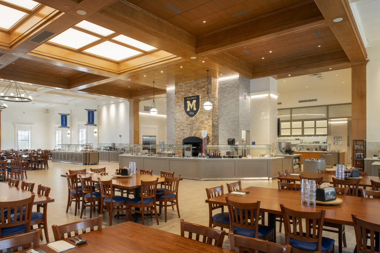 Spacious dining hall with wooden tables and chairs, high ceiling with warm lighting, and a central serving area featuring a decorative M emblem on a stone wall.