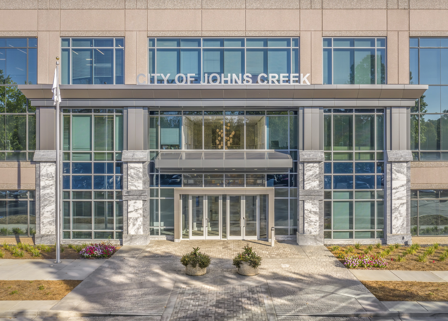 Modern two-story building with large glass windows and "City of Johns Creek" signage above the entrance, flanked by two flagpoles and landscaped planters.