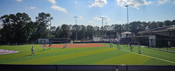 Dellinger Park Softball Complex. Youth lacrosse players compete on a striped green-and-orange field under a bright blue sky, with trees, bleachers, and stadium lights behind them.
