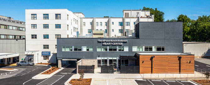 Caldwell University of North Carolina (UNC) Health Care. A modern hospital complex with white and gray buildings stands beside a marked parking lot under a clear blue sky; the entrance reads “The Afshar and Andersen Heart Center,” conveying a calm, welcoming feel.